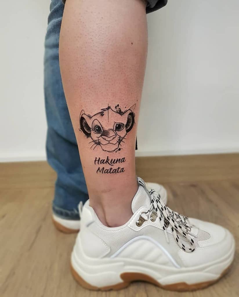 Detailed line art tattoo of Simba’s face with the phrase 'Hakuna Matata' underneath, showcasing his iconic playful expression