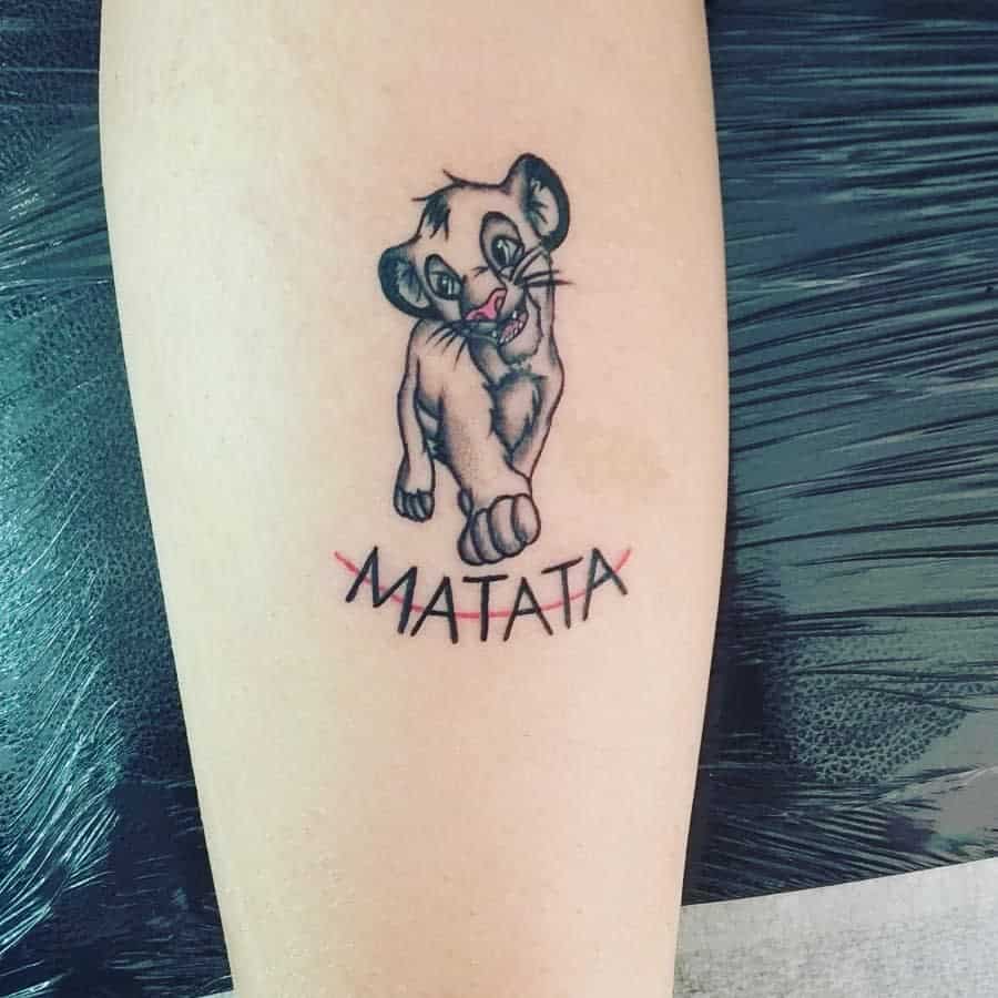 Playful tattoo of Simba walking with the word 'MATATA' crossed out below, highlighting his cheerful spirit