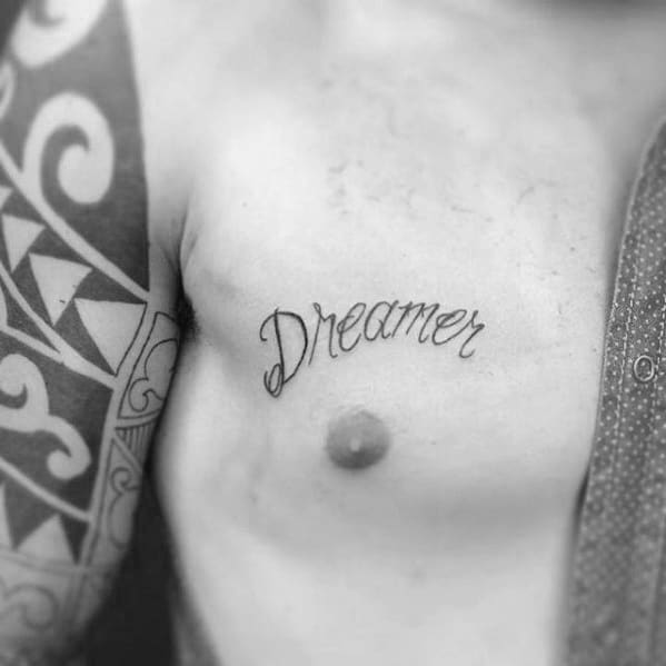 Chest tattoo with the word “Dreamer” in soft cursive script, placed above the left side of the chest near tribal-style arm ink.