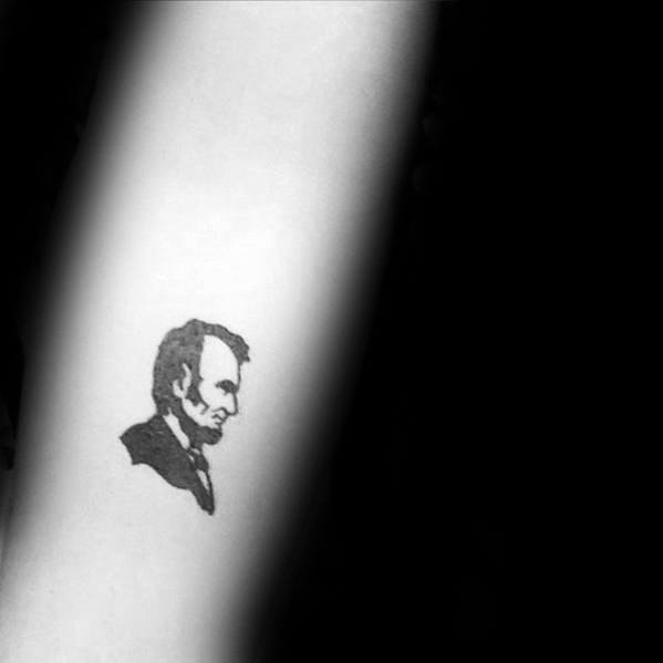 Tiny tattoo of Abraham Lincoln's silhouette on an arm, in black ink, against a dark background