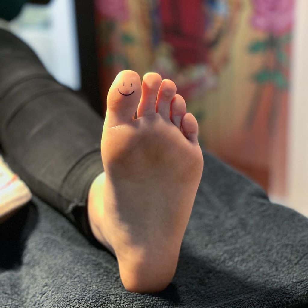 A small minimalist tattoo of a simple smiley face on the tip of a toe, creating a playful and subtle design on the sole of the foot.