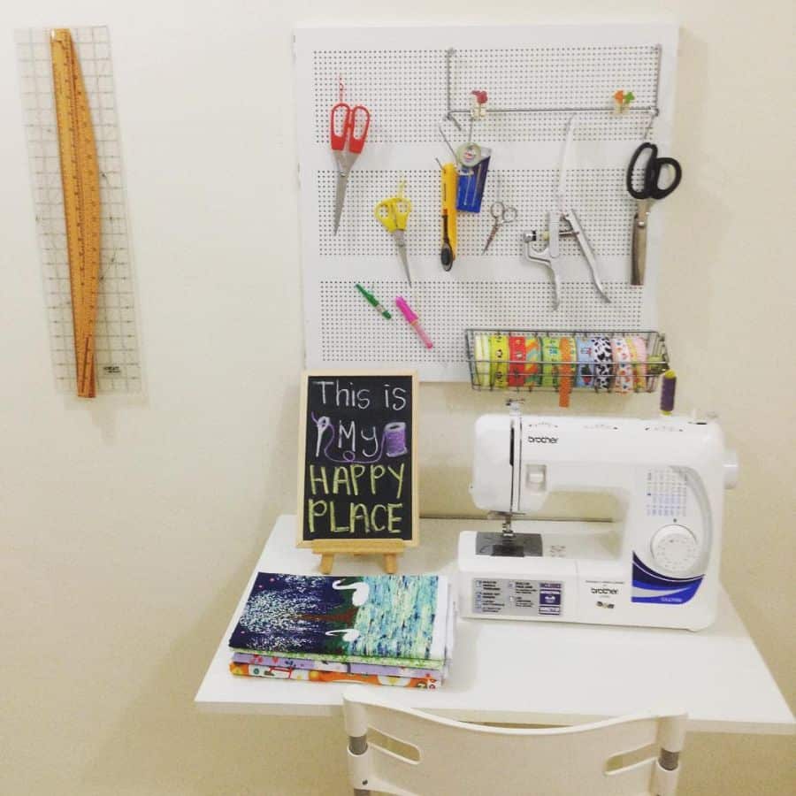 Compact sewing corner with a machine, pegboard for tools, fabric stack, and a chalkboard saying 'This is my happy place'