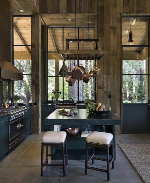 Rustic kitchen featuring wood-paneled walls, tall ceilings, and a hanging pot rack over a dark green island.