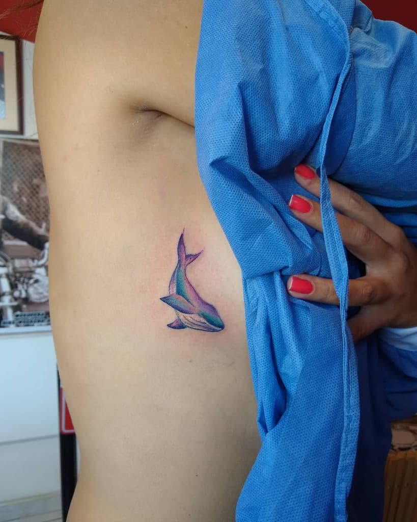 Small watercolor-style tattoo of a vibrant blue and purple whale, blending soft shading with a minimalist design