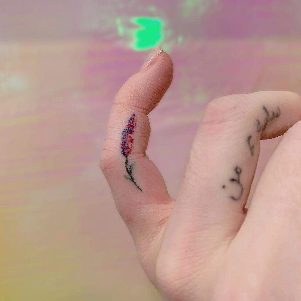 Delicate finger tattoo of a small, colorful lavender sprig in fine-line style, placed along the side of the thumb with soft shading