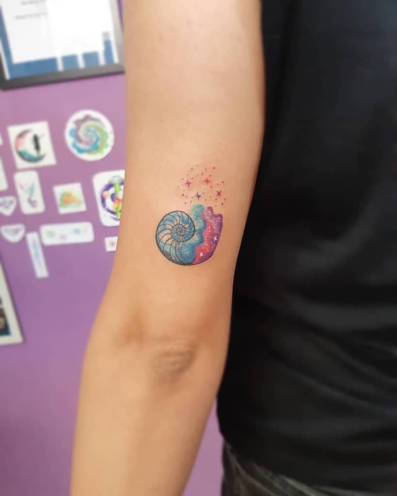 Colorful tattoo of a nautilus shell blending into a cosmic galaxy with blue, pink, and purple hues, surrounded by tiny stars