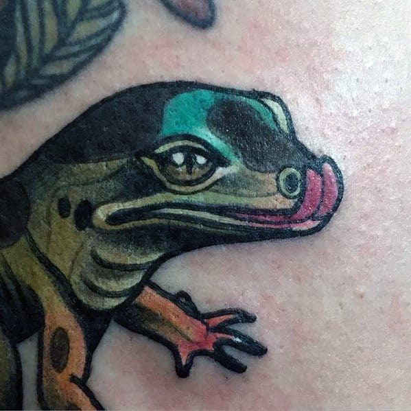Colorful gecko tattoo with bold outlines, teal and black head, and pink tongue sticking out in a playful, cartoon-style design.