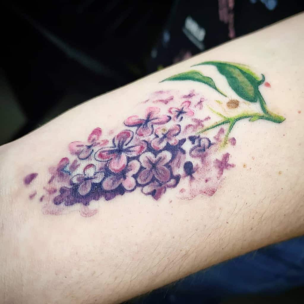 Watercolor-style tattoo of a lilac branch with soft purple and pink flowers, blending into the skin with a dreamy, artistic effect.