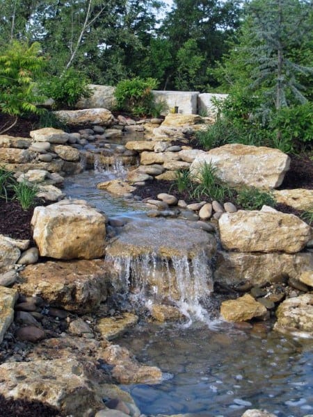 Small backyard waterfall with natural stones and a stream surrounded by greenery.