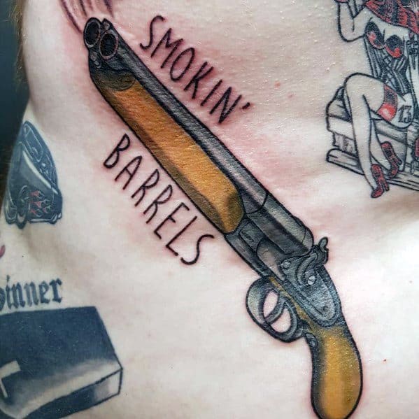 Tattoo of a double-barrel shotgun with "Smokin' Barrels" text, featuring bold black outlines, wood grain details, and dynamic shading for a classic look.