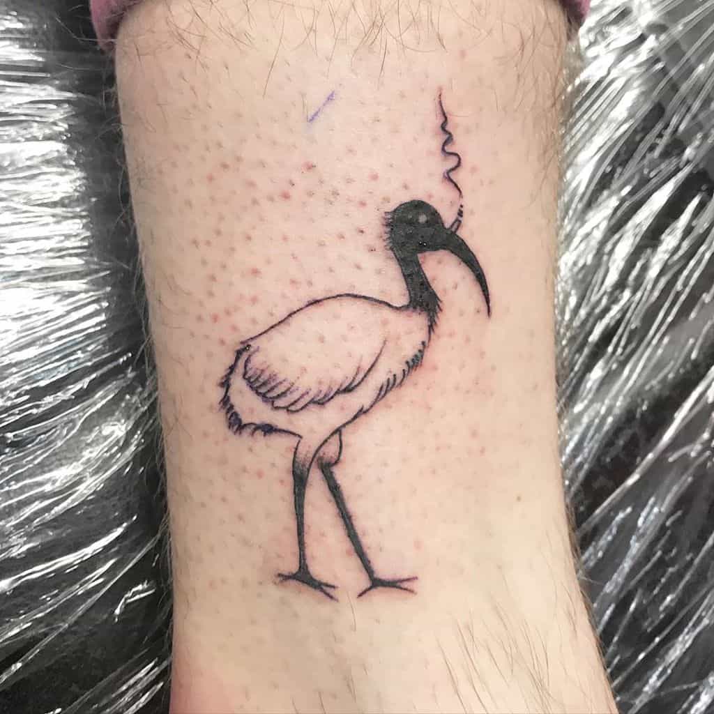 A black ink tattoo of an ibis standing with a cigarette in its beak, emitting smoke, drawn in a humorous and minimalist style on the ankle.