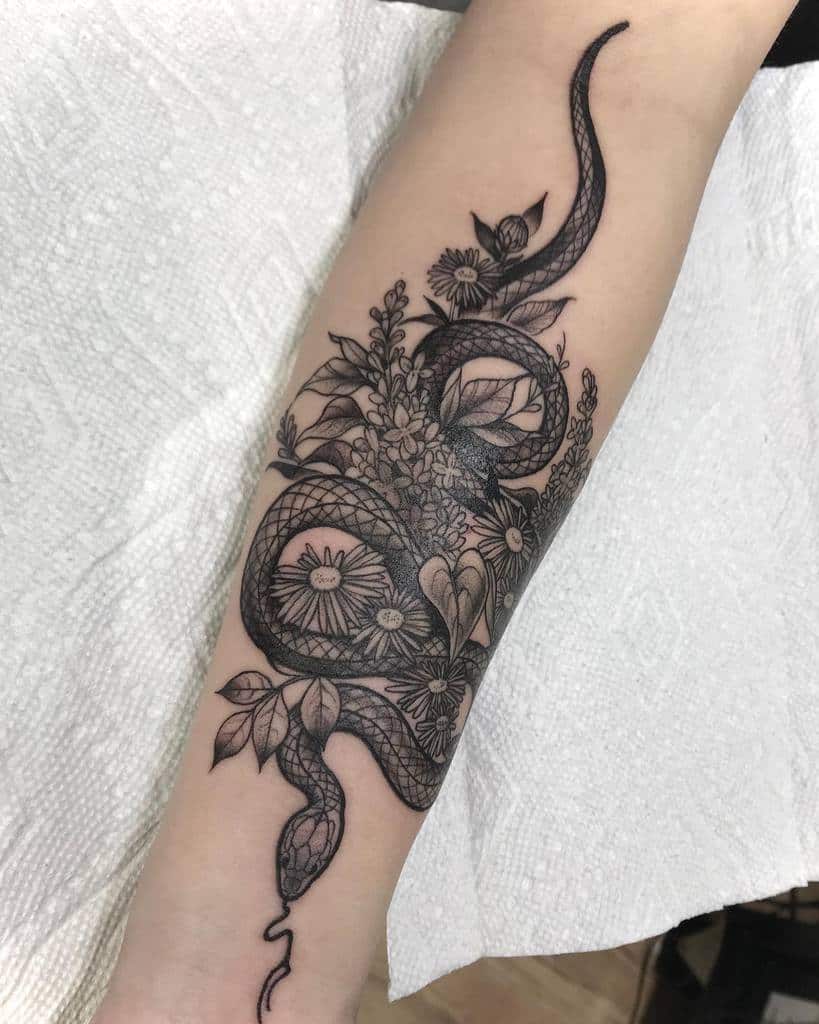 Intricate black and grey tattoo of a coiled snake intertwined with lilac flowers, daisies, and leaves, featuring fine-line detailing.