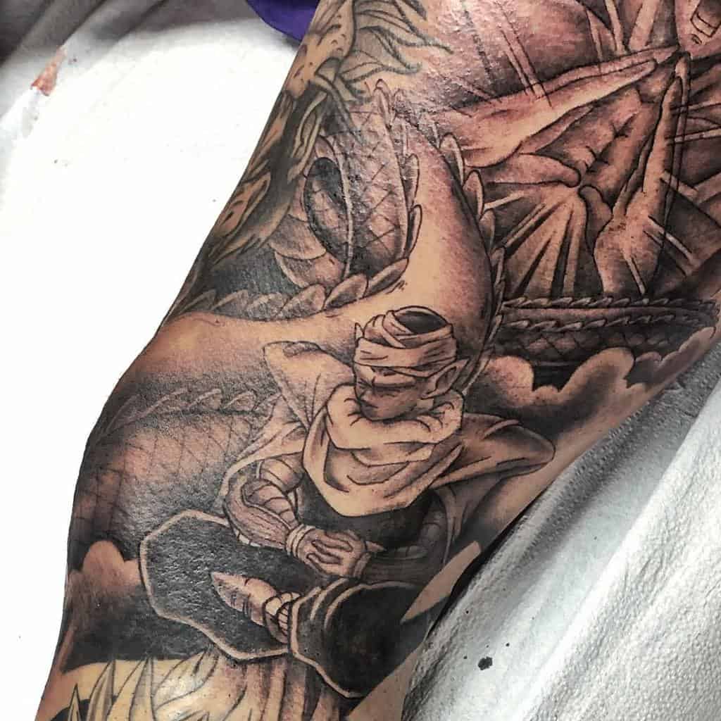 A black-and-grey tattoo of Piccolo meditating, surrounded by dramatic shading and dynamic background elements from the *Dragon Ball* universe.