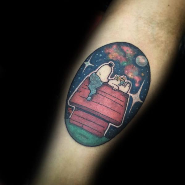 Colorful tattoo of Snoopy and Woodstock lying on a red doghouse, gazing at a starry night sky with a glowing moon, placed on an arm.