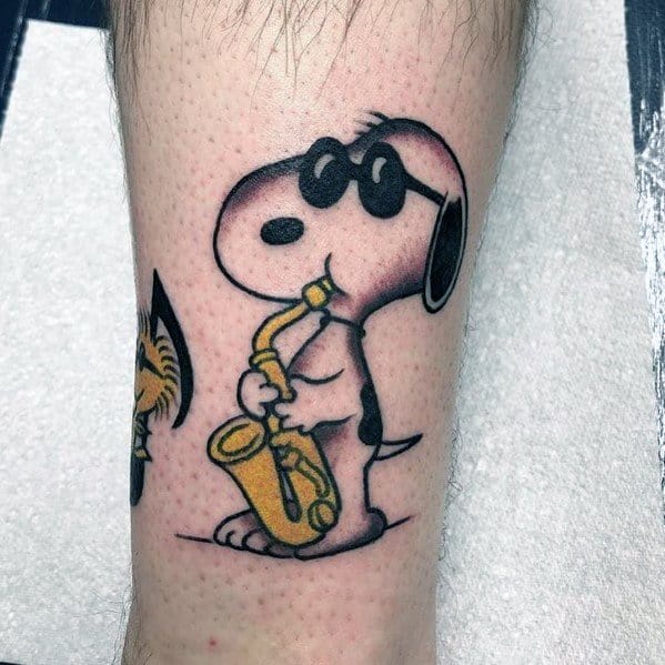Colorful tattoo of Snoopy wearing sunglasses and playing a yellow saxophone, placed on a person's leg.