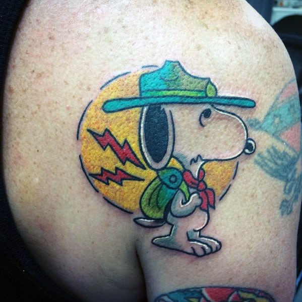 Colorful tattoo of Snoopy dressed as a scout with a green hat, backpack, and red scarf, standing against a yellow circular background, placed on a shoulder.
