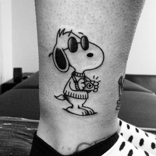 Black and white tattoo of Snoopy wearing sunglasses and a sweater, holding a small camera, placed on a person's ankle.