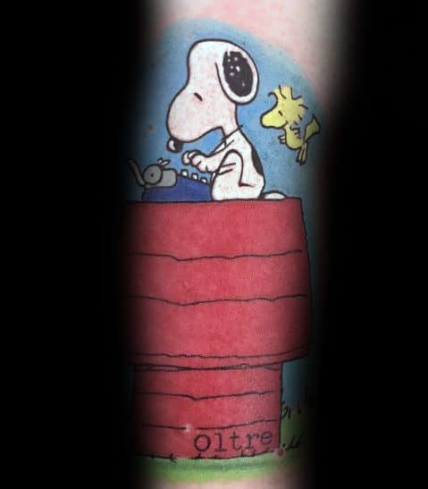 Colorful tattoo of Snoopy typing on a typewriter atop his red doghouse, with Woodstock flying nearby, set against a blue sky background.