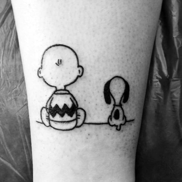 Black and white tattoo of Charlie Brown and Snoopy sitting side by side on a thin line, facing away, placed on a person's leg.