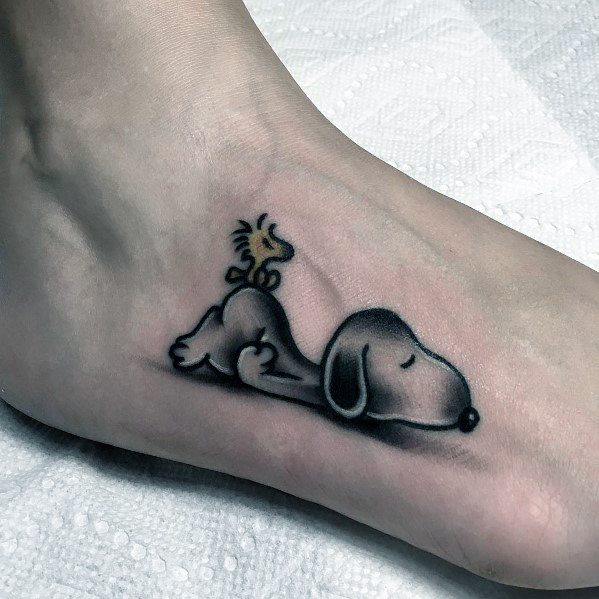 Tattoo of Snoopy sleeping on a foot with Woodstock sitting on his back, shaded in black and gray for a soft, relaxed look.