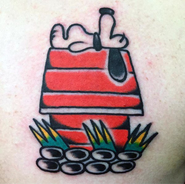 Colorful tattoo of Snoopy lying on top of his red doghouse, surrounded by grass and stones, placed on a person's skin.