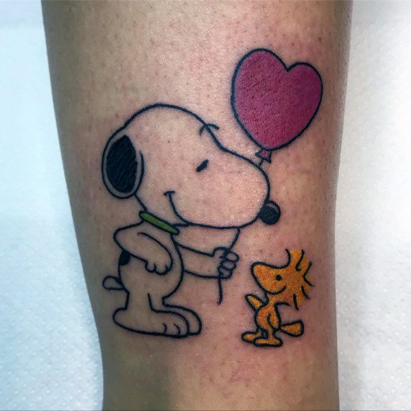 Colorful tattoo of Snoopy giving Woodstock a heart-shaped balloon, with both smiling, placed on a person's leg.