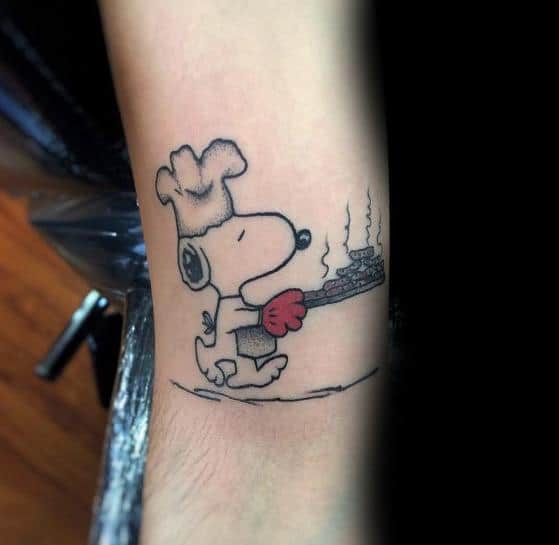 Black and white tattoo of Snoopy dressed as a chef, wearing a hat and oven mitt, carrying a steaming tray of food, placed on a person's arm.