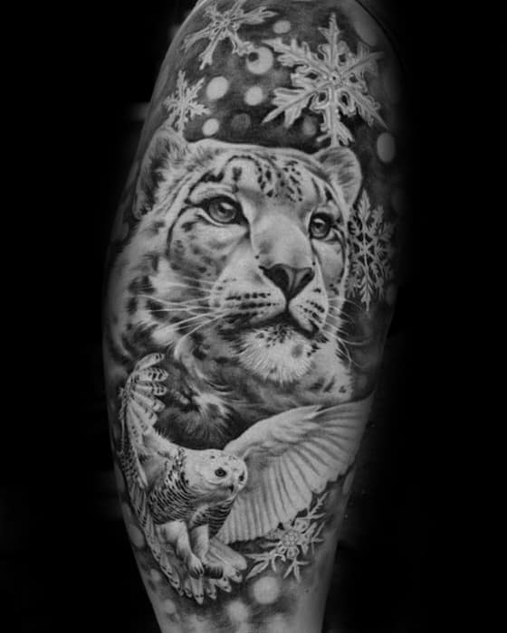 Detailed black and gray snow leopard tattoo with a soaring owl and snowflakes, blending realism and winter-themed elements on the arm.