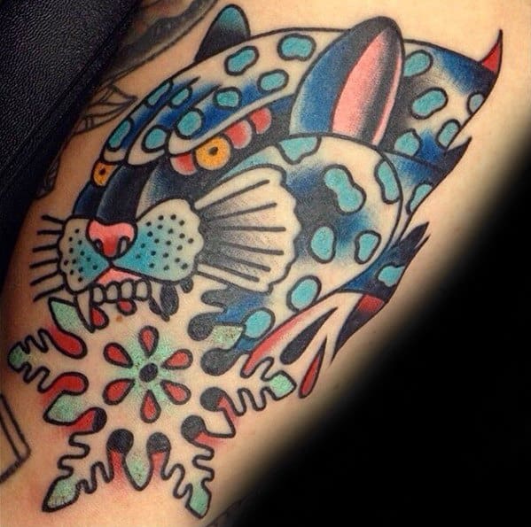 Bold neo-traditional snow leopard tattoo in blue tones, with stylized spots, intense yellow eyes, and abstract floral snowflake details.