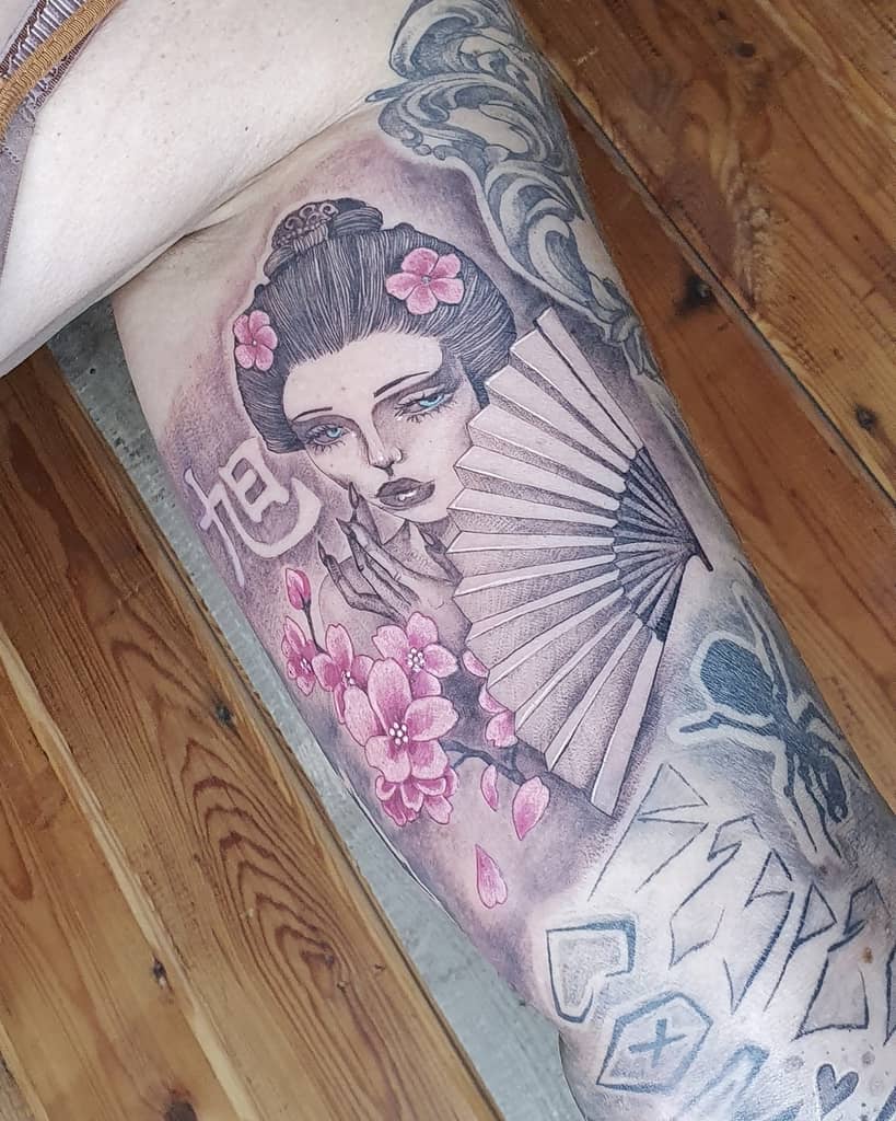 Detailed tattoo of a Geisha with piercing blue eyes, holding a fan, surrounded by cherry blossoms, kanji symbols, and flowing designs.
