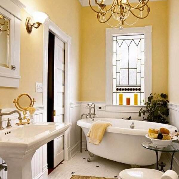 Cozy vintage bathroom with soft yellow walls, white wainscoting, chandelier, and stained-glass window.