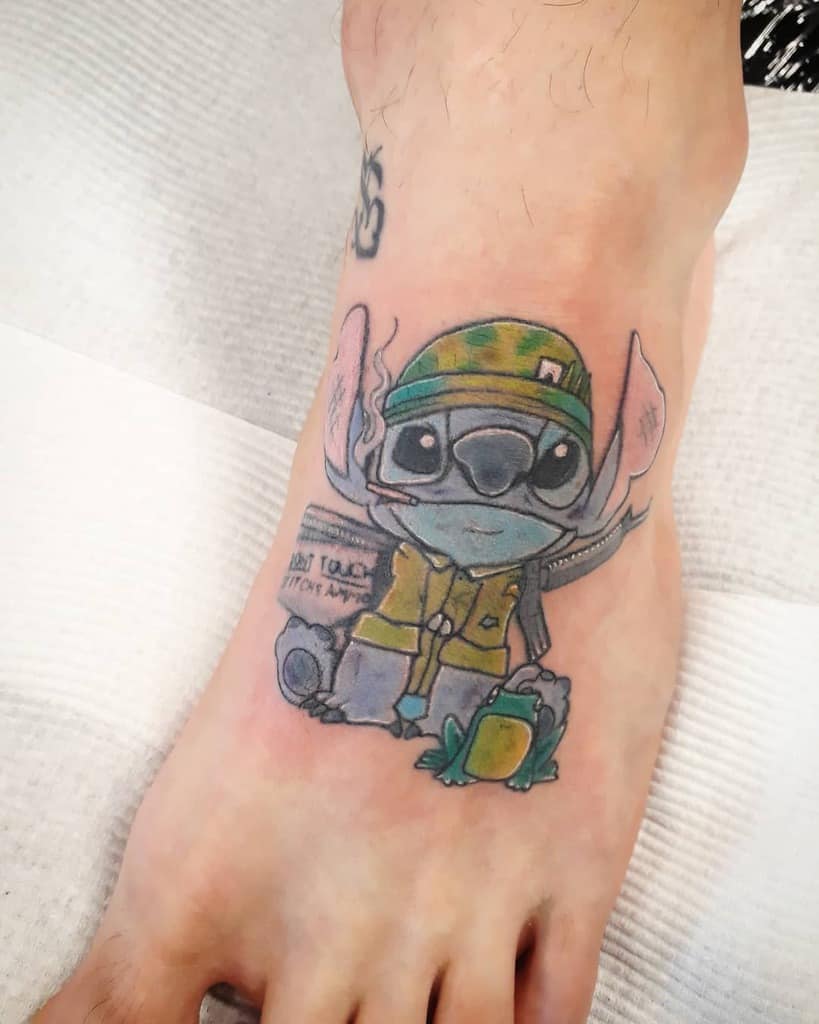 Colorful tattoo of Stitch from Lilo & Stitch wearing a military helmet and uniform, holding a frog, on the foot.