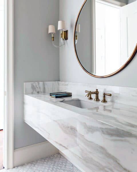 Solid marble vanity with brass faucet, round mirror, and wall sconces.