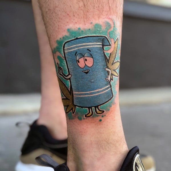 South Park tattoo of Towelie holding a joint, with a smoky blue-green background and cannabis leaves, featuring bold colors and shading.