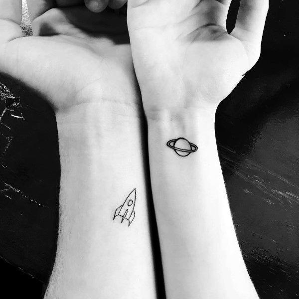 Minimalist matching wrist tattoos of a rocket and a planet with rings, symbolizing a love that’s adventurous, boundless, and written in the stars.
