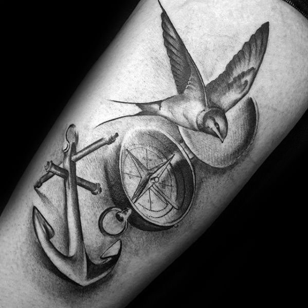 Tattoo of a bird, anchor, and compass on a person's arm, all in black and grey tones, symbolizing guidance and stability