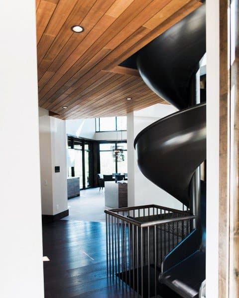 The modern home interior features a sleek black spiral slide, wooden ceiling, and expansive windows in the living area