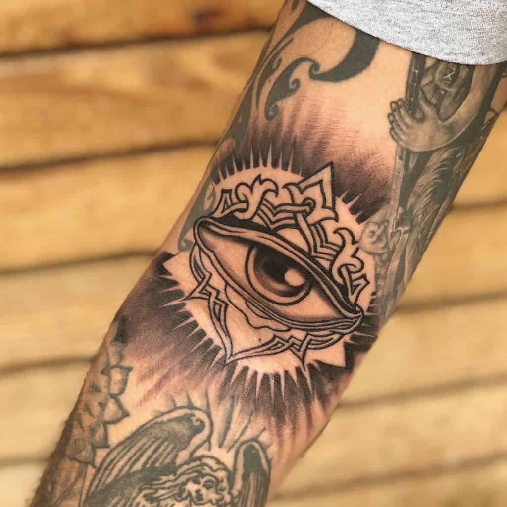 Tattoo of a realistic all-seeing eye inside an ornate design with radiating lines, inked in black and grey shading on the forearm.