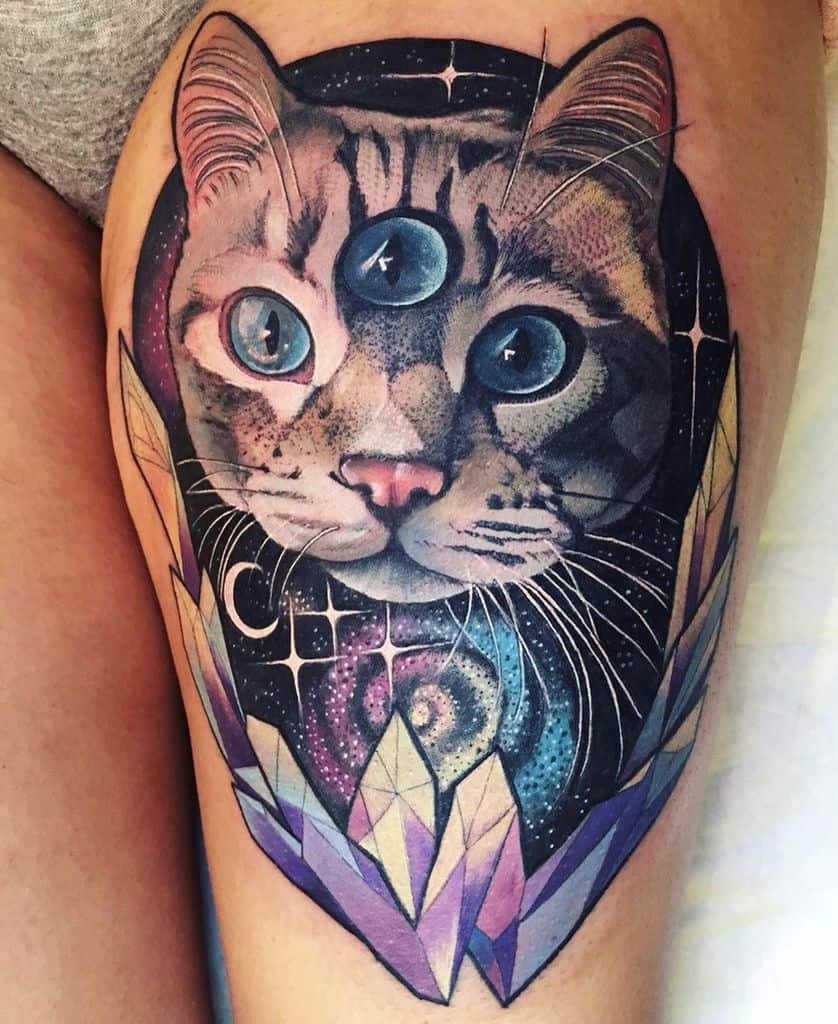 Tattoo of a cosmic three-eyed cat with glowing blue eyes, surrounded by a galaxy background and crystal formations, inked on the thigh.