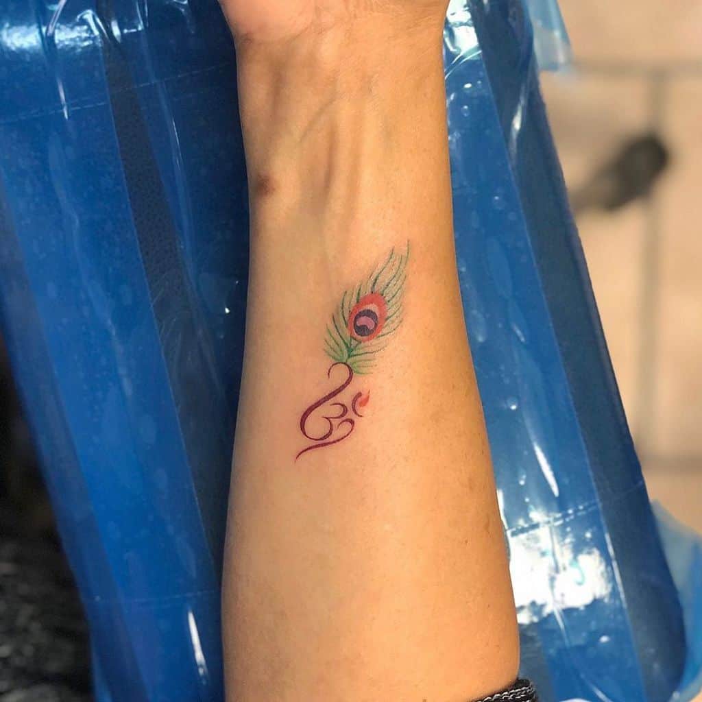 Small, delicate peacock feather tattoo on the wrist, featuring fine green and blue hues with an 'Om' symbol in red ink for a spiritual touch.