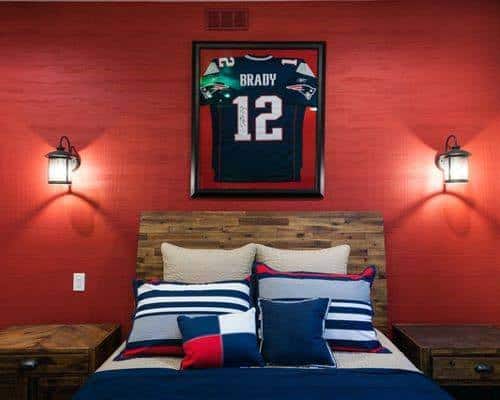 Bedroom with a wooden bed, red-striped pillows, and a framed sports jersey on a red wall lit by two wall lamps