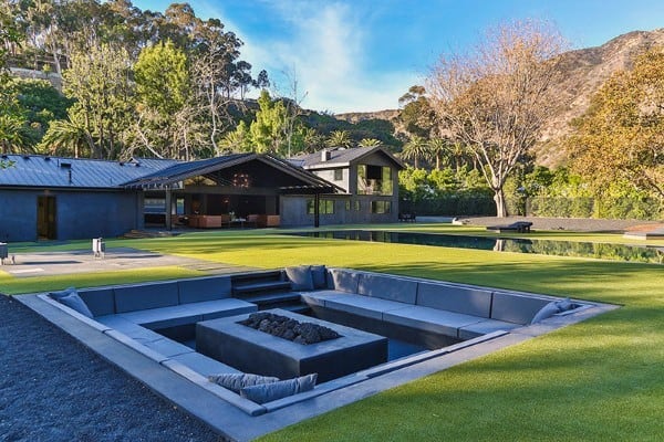 Cozy sunken seating area with a fire pit, modern house, lush lawn, and hillside trees in the background