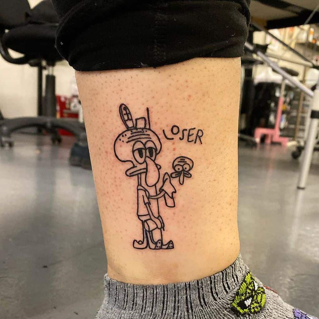 A black ink tattoo of a grumpy cartoon squid character with 'LOSER' written above, drawn in a humorous and minimalist style on the lower leg.
