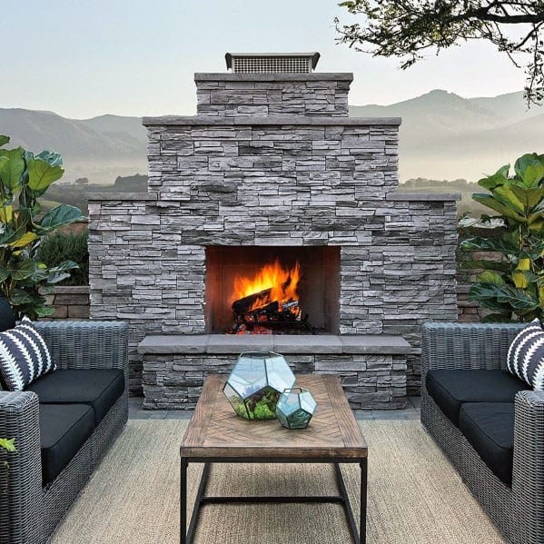 Stacked stone fireplace with a lit fire, seating, and plants, set against mountain views