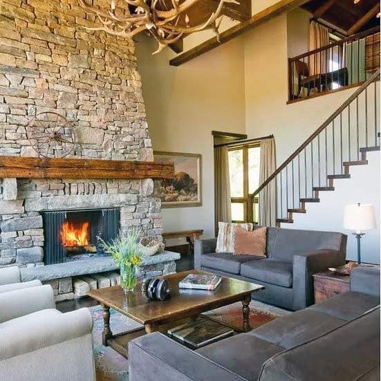 Rustic living room with a stacked stone fireplace, wooden mantel, antler chandelier, and cozy seating.