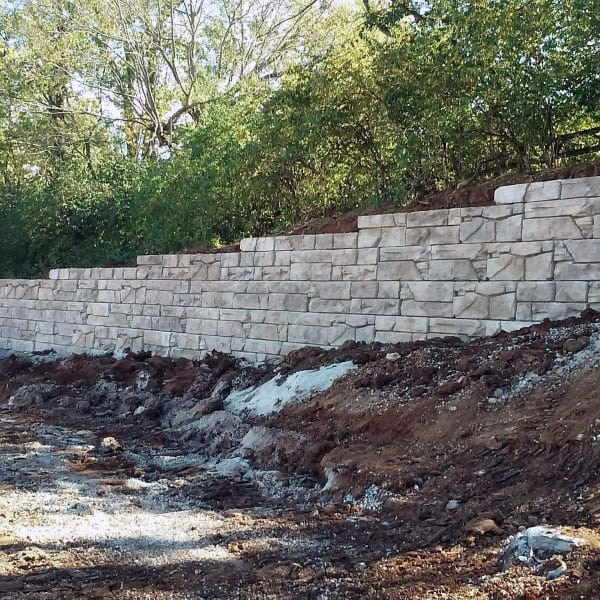 Stacked stone retaining wall under construction with surrounding soil and greenery.