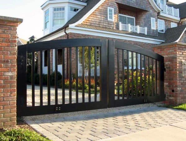 Large black metal gate in front of a brick driveway leading to a house with a sloped roof and multiple windows