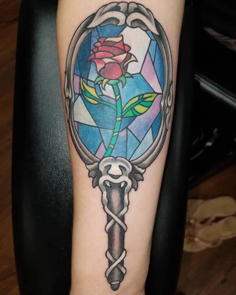 A stained-glass style Beauty and the Beast rose tattoo inside a dome, framed by an ornate mirror with vibrant, colorful details