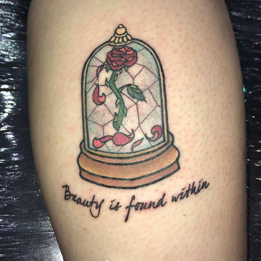 A Beauty and the Beast-inspired tattoo featuring a rose inside a stained glass dome with the quote "Beauty is found within" beneath it