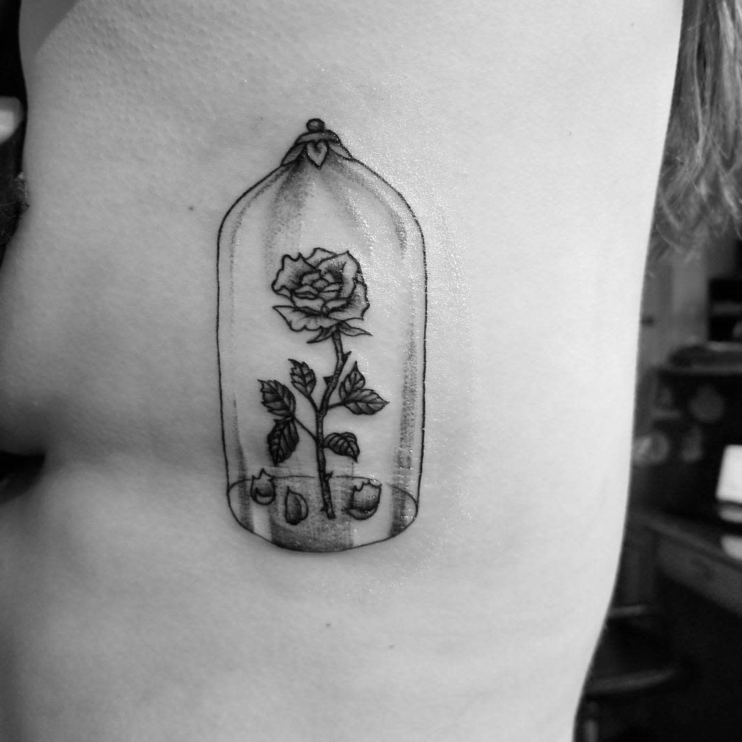A black and white Beauty and the Beast rose tattoo inside a glass dome, featuring fine details on the rose and petals